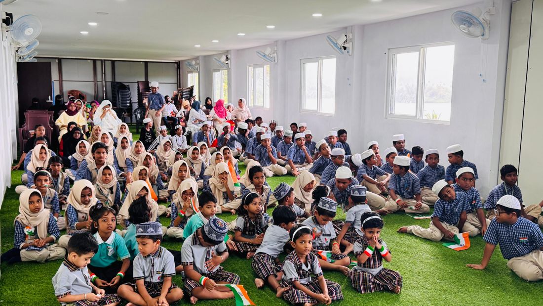 Students in uniform seated on the floor of the school hall holding small Indian flags, with parents and teachers seated behind
