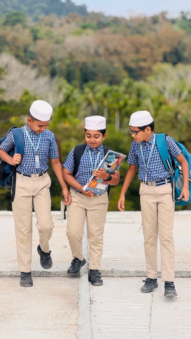 Three boys in school uniform with backpacks walking together and looking at a book, with green hills behind